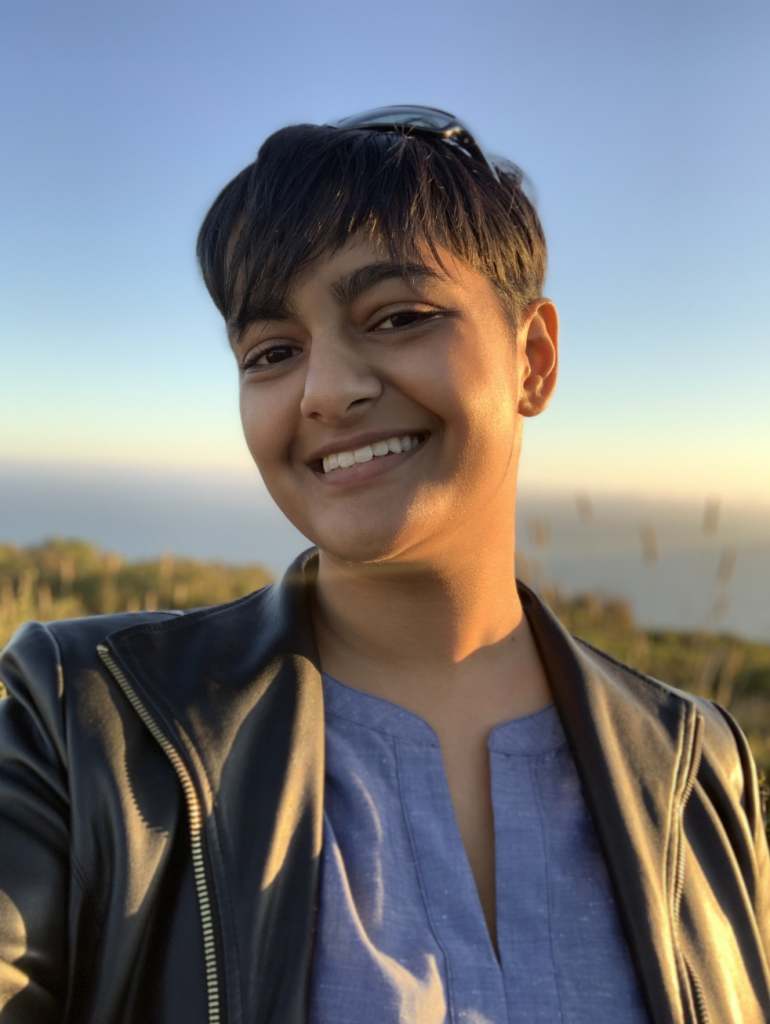 Image depicts Jay Shreedhar, a Tamil-American trans man. He has medium brown skin, short black hair in a fringe, and brown eyes. He is smiling at the camera and wearing a blue shirt with a black leather jacket. The setting sun lights up the left side of his face and the ocean is visible behind him.