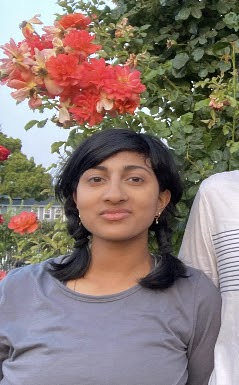 Alt Text: A picture of Meghna Chandrasekar, an Indian-American woman. She has medium brown skin, dark brown eyes, and black hair in two braids. She is wearing a gray shirt and standing against a background of red flowers and green leaves.