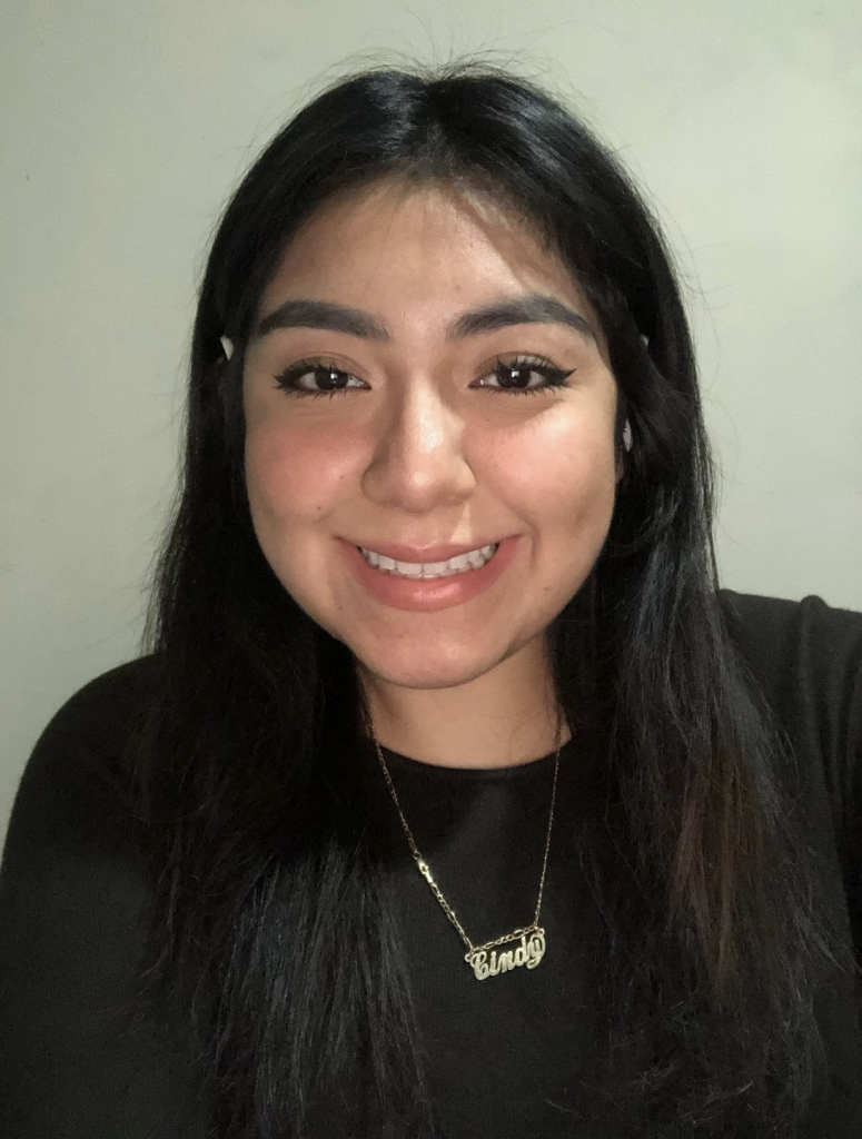 Alt Text: A picture of Cindy Esperanza Sanchez, a Mexican-Salvadoran-American woman. She has long black straight hair, dark brown eyes, thick eyebrows, and light brown skin. There is a gold chain with her name [Cindy] in cursive. They are wearing a fitted, knitted black long sleeve shirt. They are wearing white, noise canceling earphones. They are smiling and are against a white, blank backdrop.