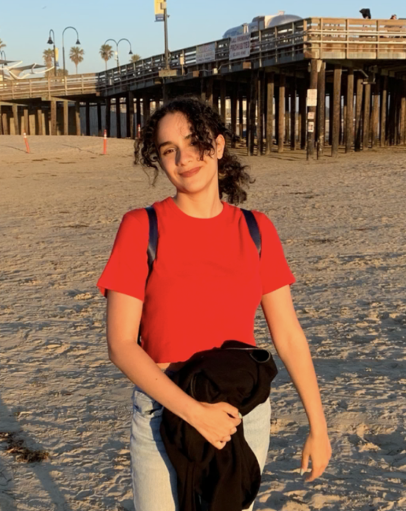 A picture of Yasmine Hirech, a light-skinned woman with dark brown curly hair in a ponytail. Some of the curls frame her face. She is wearing a red shirt and pale blue jeans and holding a black jacket while smiling at the camera. Behind her is a backdrop of a beach boardwalk.